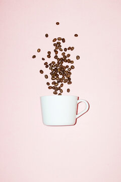 Roasted coffee beans fall into a white cup on a pink background