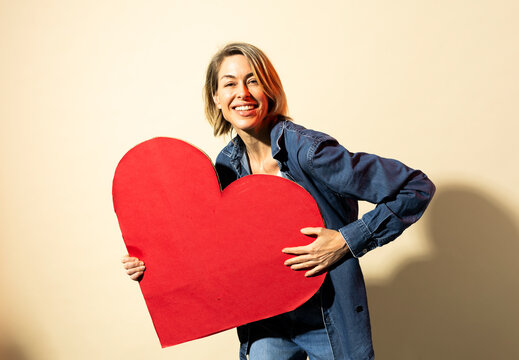 Smiling woman holds a red paper heart indoors showing love
