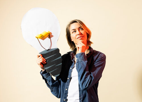Thoughtful woman holds a paper light bulb while brainstorming indoors