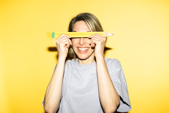 Smiling woman holding oversized pencil over eyes on yellow background