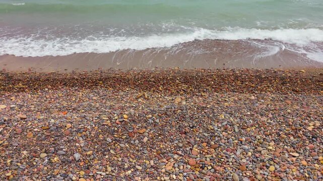 Colorful pebble beach beside foamy sea water from elevated coastal view. Small waves approaching stony shore. Tranquil marine landscape with wet stones, surf patterns and coastal nature