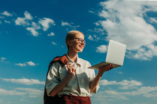 Smiling businesswoman holds a laptop outdoors under a blue sky