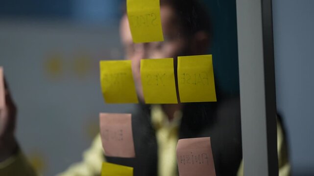 A designer in a yellow shirt behind glass reaches up to place a note among the cards already stuck to the pane inside a dimly lit machine learning startup office.