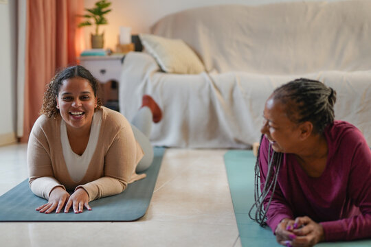 Happy curvy african daughter having fun doing yoga exercise with mother at home during fall season - Family lifestyle, wellbeing and happiness concept