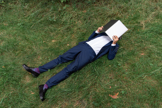 Man lying on green grass with laptop covering face, taking a wor