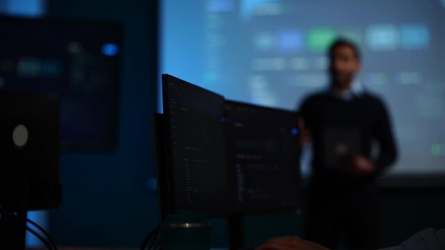 A presenter dissolved into shallow focus keeps talking behind a rank of monitors that hold the live orchestration dashboard in sharp view in a dark tech office.