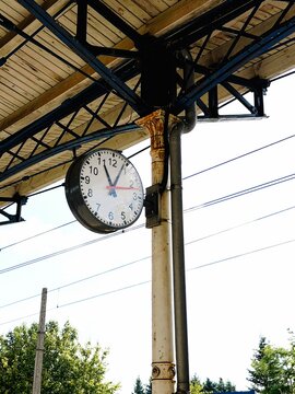 Round wall clock displaying time at a train station platform with metal beams and trees visible in the background under a clear blue sky