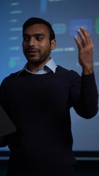 A machine learning engineer holding a tablet talks straight to camera in a long unbroken take with the agent monitoring board glowing behind him in a dark tech office.