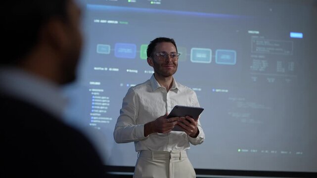 A speaker framed past a listener's shoulder works through the orchestration board at length while the foreground stays a dark silhouette inside a dark AI startup office.