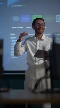 A presenter in a white shirt at the wall display holds the tablet flat and lets the room read the projected pipeline over his shoulder during a late night shift.
