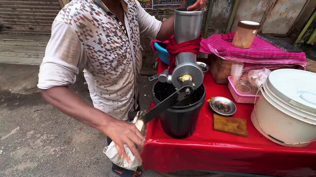 A street vendor operates a compact manual juice extraction machine, pouring fresh ingredients into the metal hopper while collecting the extracted juice in a cup, capturing an authentic Indian street 