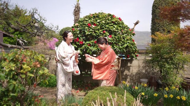 Two kimono-clad adults stand in a garden while one holds a red ball attached to a string toy.