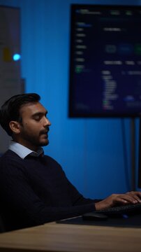 An engineer at his keyboard in half profile works on quietly with a wall of code out of focus behind his shoulder inside a dimly lit machine learning startup office.
