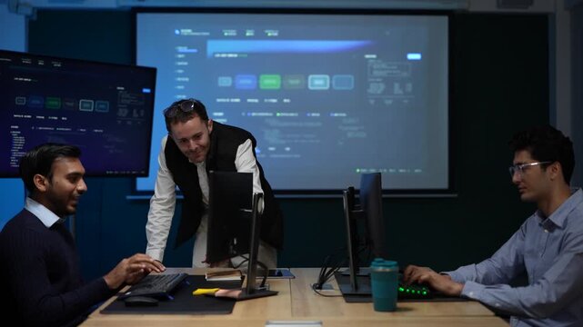 Three engineers along a workstation row share a moment of relief with the projected agent board lighting the room behind inside a dimly lit machine learning startup office.