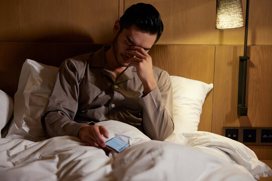 Tired man rubbing eyes while using smartphone in bed at night