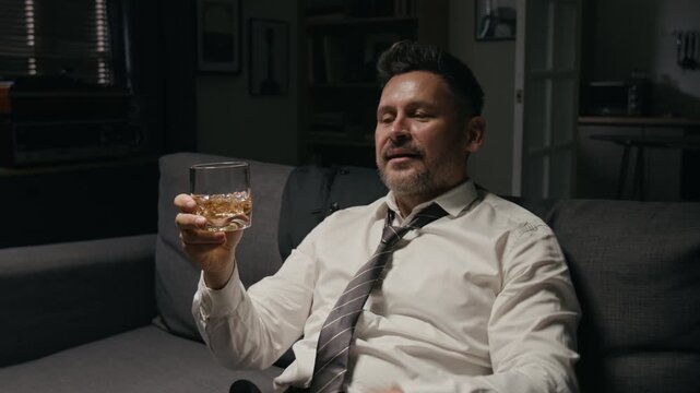 Middle aged Caucasian businessman relaxing on couch, drinking whiskey from glass after work. Clip represents alcohol abuse, burnout, stress coping and mental health awareness