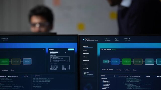 Two monitors carrying the orchestration board stay in focus while the team works the problem out of focus behind the desk inside a dimly lit machine learning startup office.