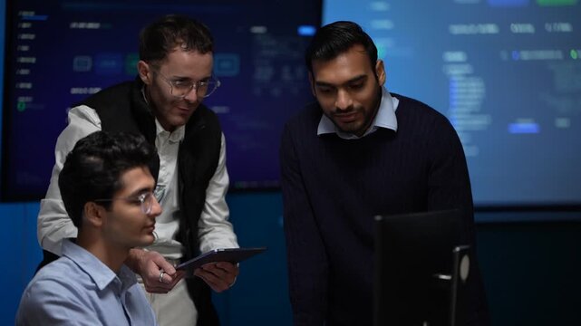 Colleagues leaning over a shared monitor point into the same panel from either side and then straighten up smiling inside a dimly lit machine learning startup office.