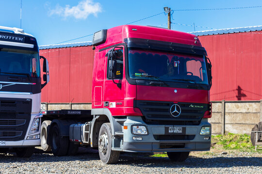A red Mercedes‑Benz Actros and a white Volvo FH tractor unit stand side by side on a gravel lot in Angol, Chile