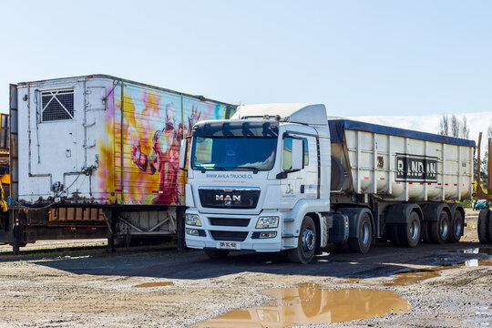 Two large trucks parked on a muddy lot in Angol, Chile, including a white semi‑truck and a vibrant graffiti‑covered trailer under clear daylight