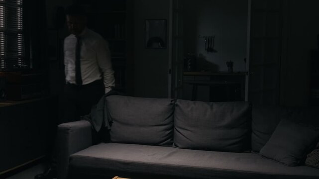 Exhausted middle aged Caucasian businessman collapsing onto couch and loosening tie in dark room. Ideal visual for workplace stress, burnout and mental health projects