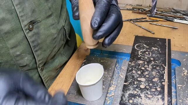 Gloved artisan applying liquid adhesive from a small cup onto a wooden handle, inserting a forged metal spoon component, and securing the assembly with a small metal pin fastener on workbench