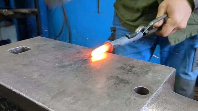 A skilled metalworker uses traditional blacksmithing techniques to shape a glowing hot iron rod on a heavy steel anvil using a hammer and tongs in an industrial workshop environment
