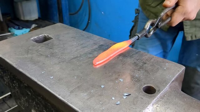 A skilled metalworker uses traditional blacksmithing techniques to shape a glowing hot iron rod on a heavy steel anvil using a hammer and tongs in an industrial workshop environment
