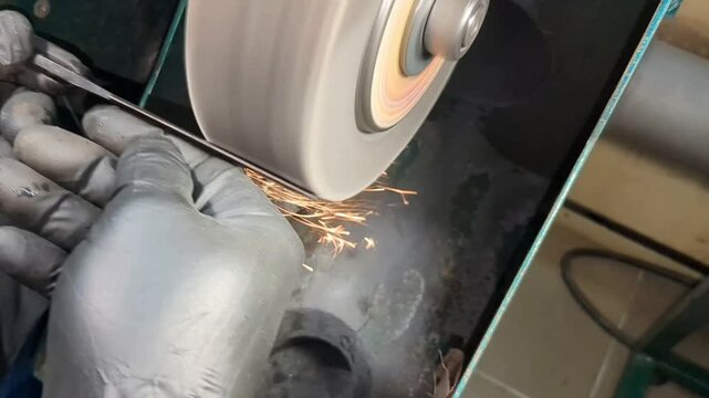 An industrial worker wearing protective black gloves operates a mechanical bench grinder to shape and smooth a metal rod, generating bright sparks during the precision manufacturing process in a works