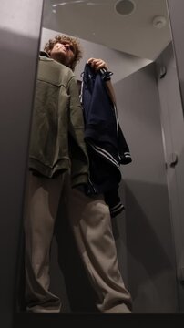 Vertical screen shopper holds two sweaters in his hands and thinks about which one to buy while comparing their style color and fit before making his final choice in shop