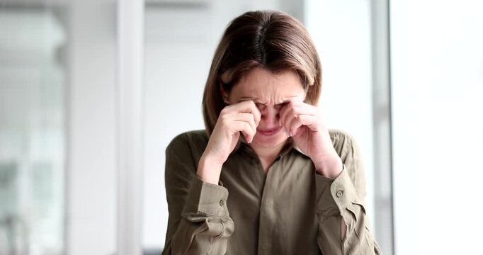 A woman sits in a calm, bright room, touching her face and eyes as she processes a wave of emotions. Her gestures convey deep introspection and vulnerability.