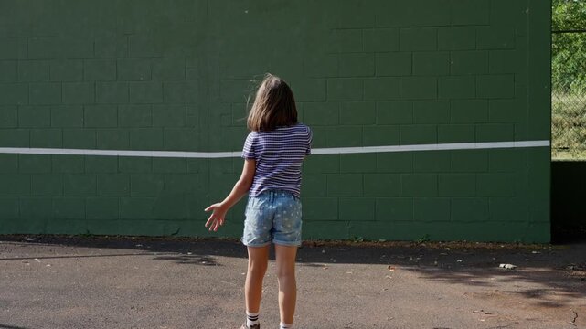 Youth practicing tennis against green cinderblock training wall with white stripe, rear viewpoint showing full swing progression and racket follow-through