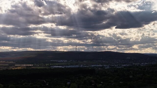 Sun rays stream through layered dramatic clouds above rolling wooded hills and a distant town, creating contrasting light and shadow across the landscape.