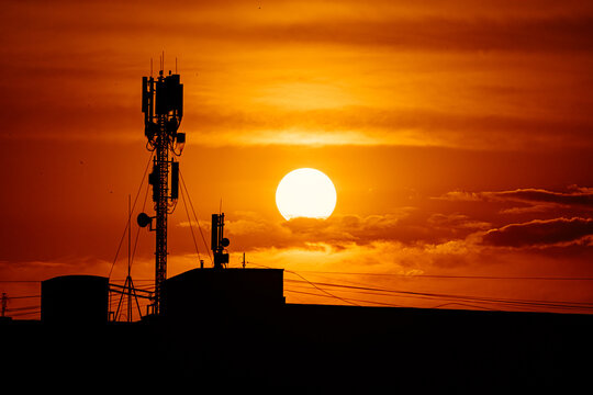 A striking silhouette of a telecommunications tower, antennas, and building rooftops against a vibrant, deep orange sunset sky.