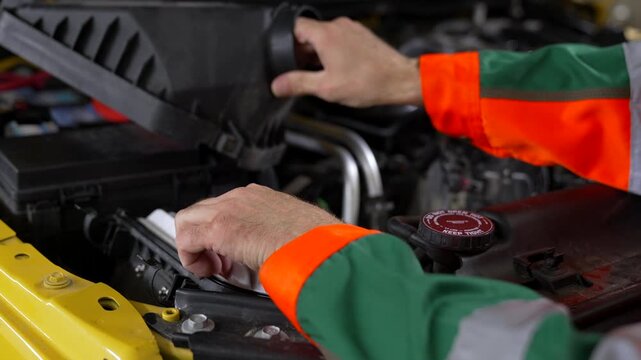 Mechanic checks engine air filter. Hands steady the new filter. Work gloves are bright orange and green. Engine bay is open, tools nearby. Perfect for auto repair or maintenance content