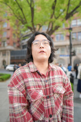 Fototapeta premium A vertical outdoor portrait shows a person in a plaid shirt looking upward beneath green street trees.
