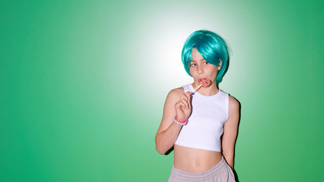Young girl with bright blue wig and white crop top eating a lollipop against a vibrant green wall