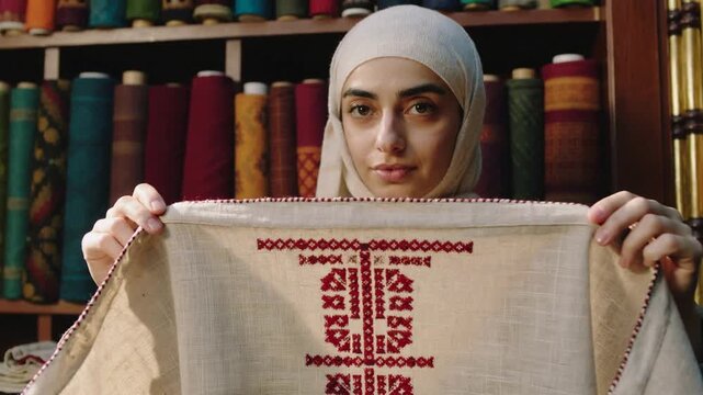 Noticing hem, artisan in beige hijab pinching, unfolding cloth at shop, revealing red embroidery