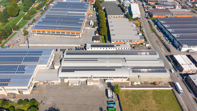 Aerial view of an industrial zone featuring large warehouse buildings with solar panels on their roofs, parking areas, and surrounding roads in Sesto Ulteriano, Lombardy, Italy.