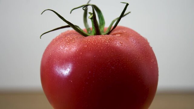 A ripe red tomato with droplets slowly rotates against a light background