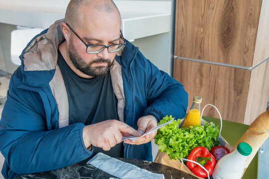 Concerned man check grocery receipt while sit in supermarket with shopping bag filled with food products. Daily expenses, smart budgeting, saving money, price awareness, personal finance