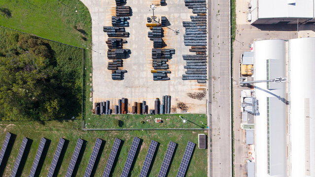 Aerial view of a metal construction company facility with organized rows of steel pipes in an outdoor yard, adjacent to an industrial building and solar panels in Cornaredo, Lombardy, Italy.