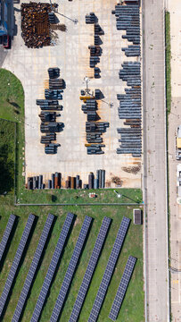 Aerial view of a metal construction company storage yard with stacked steel pipes and rows of solar panels on the grass in Cornaredo, Lombardy, Italy.