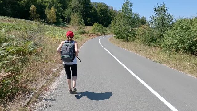 Fit senior woman carrying a backpack, in great shape and excellent physical condition, walking on the scenic mountain road. Concept of active aging and healthy lifestyle. 