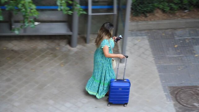 Turista viajera con maleta azul, viajera con tel&eacute;fono m&oacute;vil turista con celular y blue suitcase caminando por calle y acera de adoqu&iacute;n al aeropuerto de vacaciones de verano