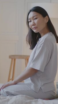 Slow motion portrait of beautiful Asian woman in pajamas sitting on bed in stylish bedroom and looking at camera. House and people concept.
