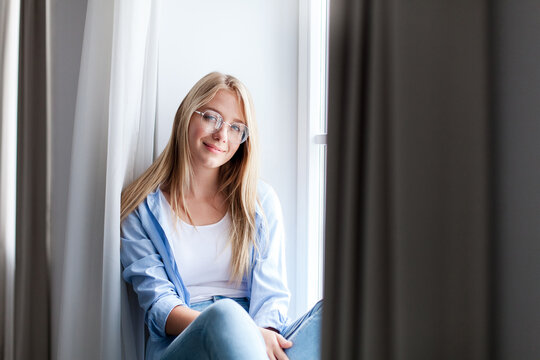 Relaxed young woman sitting on windowsill at home. Happy girl enjoying slow living. Concept of female mental health, social rest, mindfulness. Copy space