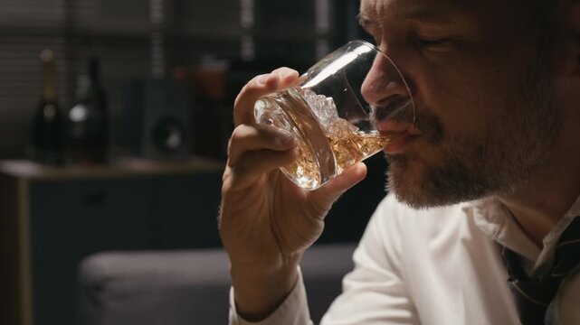Middle aged Caucasian businessman swirling whiskey in glass and drinking alcohol. Footage suits addiction recovery campaigns, corporate burnout content, and mental health awareness projects