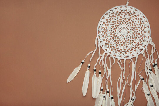 Handmade dream catcher hanging on a rope with feathers, threads, and beads on brown background.