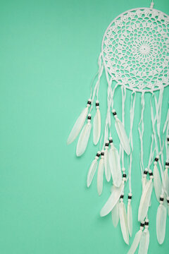 Handmade dream catcher hanging on a rope with feathers, threads, and beads on mint background.